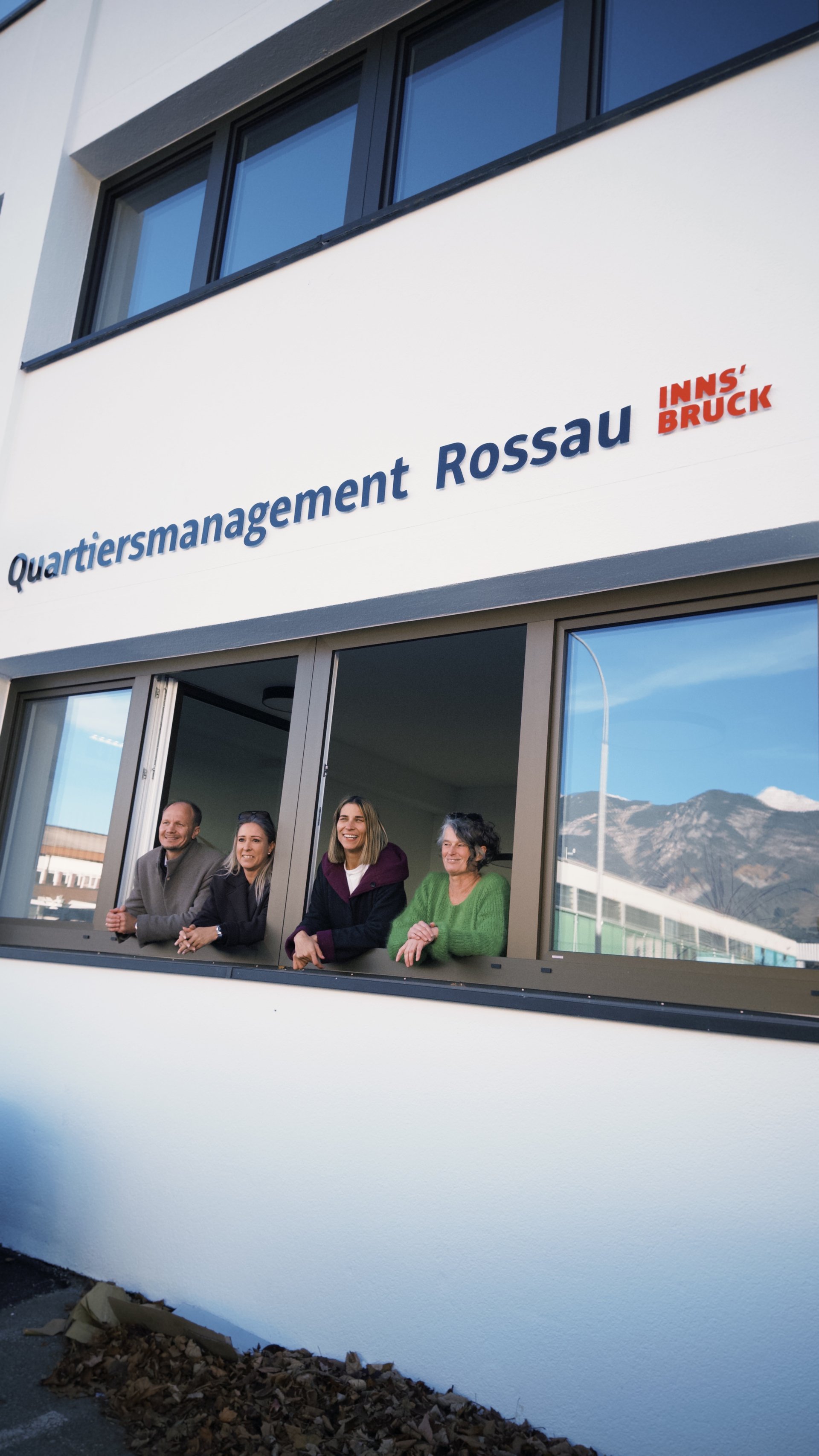 Bürgermeister Johannes Anzengruber, Stadträtin Mariella Lutz, Stadträtin Janine Bex und Quartiersmanagerin Gudrun Pechtl schauen aus dem Fenster. Über dem Fenster ist Quartiersmanagement Rossau zu lesen.
